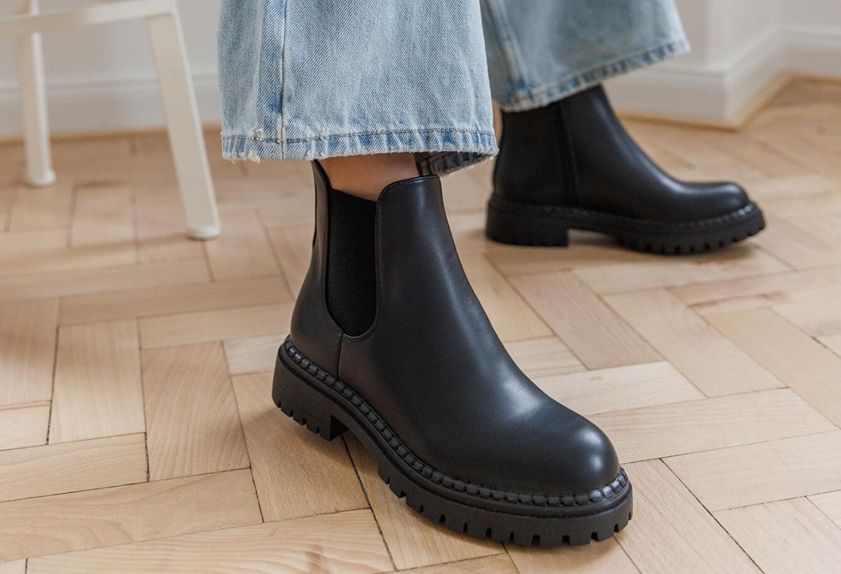 Black Chelsea boots worn with light blue jeans on a wooden floor.