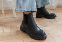 Black Chelsea boots worn with light blue jeans on a wooden floor.