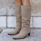 Beige knee-high boots worn by a person standing on a stone pavement with a stone wall background.