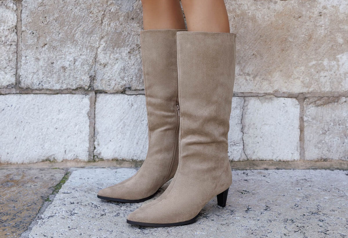 Beige knee-high boots worn by a person standing on a stone pavement with a stone wall background.