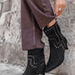 Black studded ankle boots worn with a long brown coat against a stone wall.