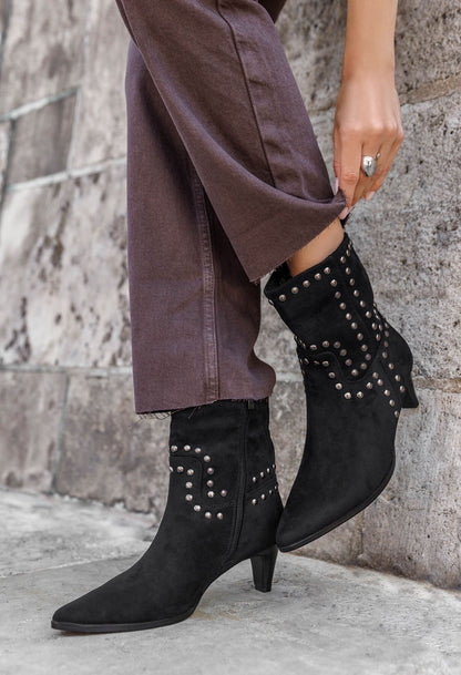 Black studded ankle boots worn with a long brown coat against a stone wall.