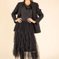 Woman wearing a black outfit with a jacket, shirt, and tulle skirt on a beige background