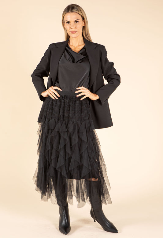 Woman wearing a black outfit with a jacket, shirt, and tulle skirt on a beige background