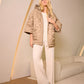 Woman wearing a beige quilted jacket and white pants in a minimalistic room.