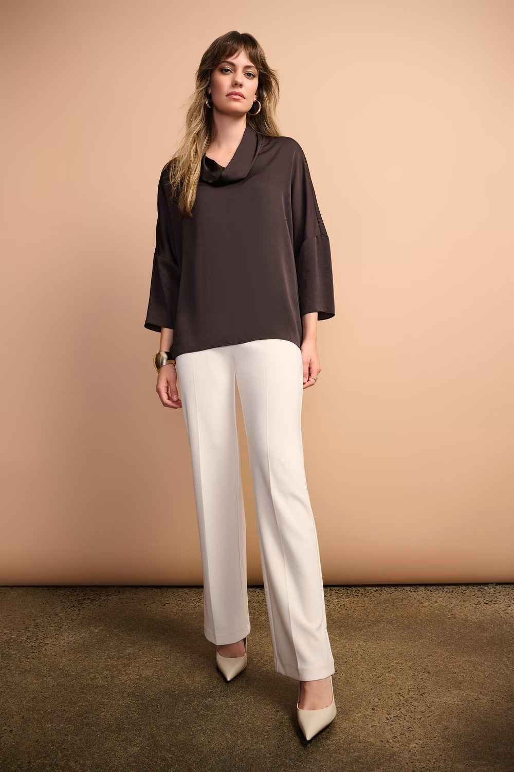 Woman wearing a dark brown top and white pants against a beige background