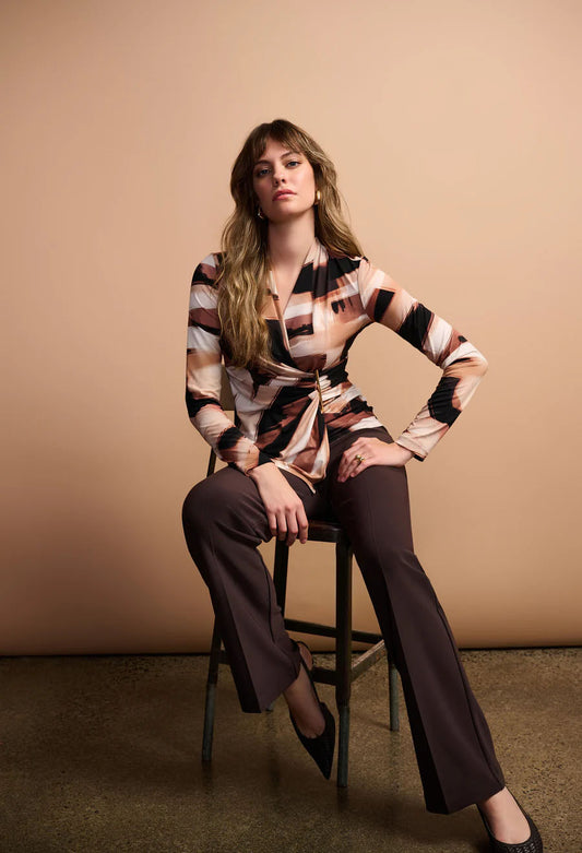 Woman sitting on a stool wearing a patterned shirt and brown pants against a beige background