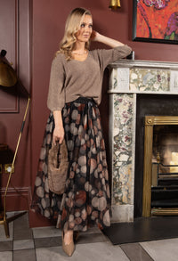 Woman in a patterned skirt and beige sweater standing in a room with a fireplace.