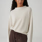 Woman wearing a white textured sweater and brown pants on a plain background