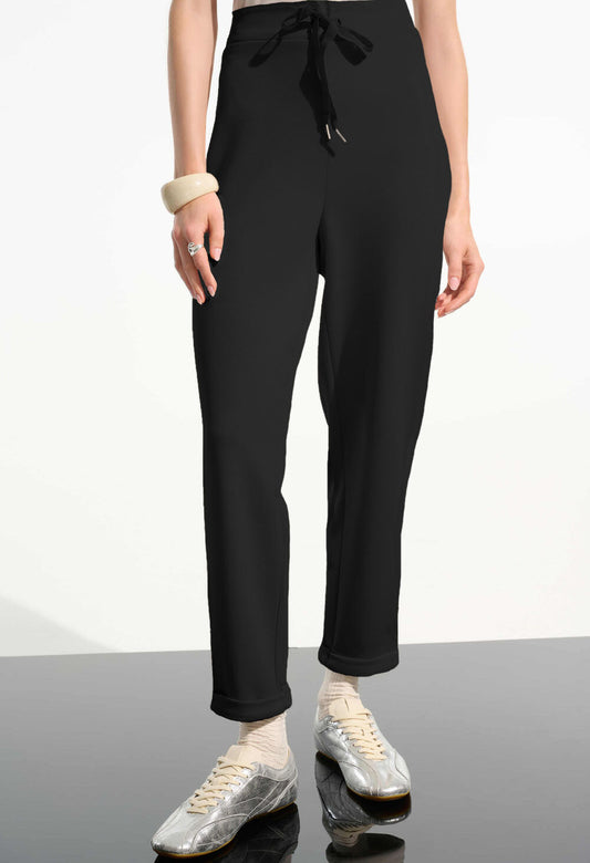 Person wearing black high-waisted pants with a tie and silver shoes on a reflective surface.