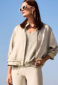 Woman wearing a beige jacket, gold top, and white pants against a blue sky.