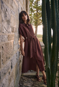 Woman in a brown dress standing against a stone wall with cacti in the foreground