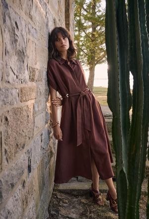 Woman in a brown dress standing against a stone wall with cacti in the foreground