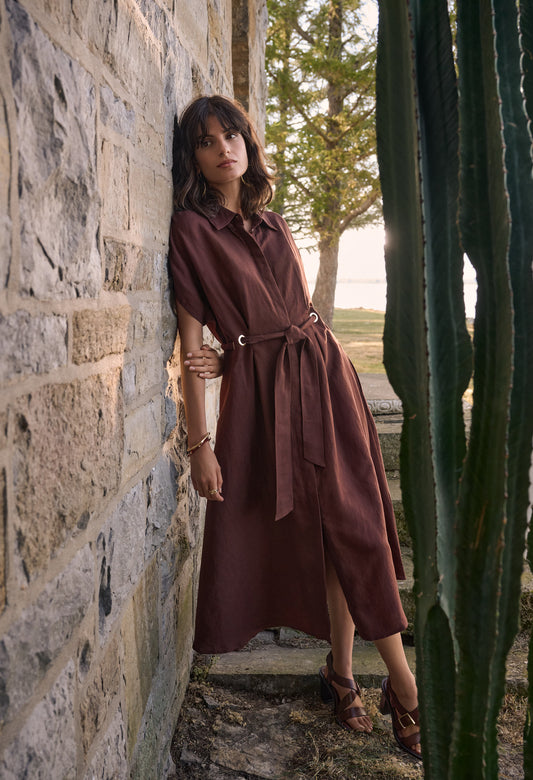 Woman in a brown dress standing against a stone wall with cacti in the foreground