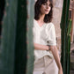 Woman in a white sweater and beige pants standing between two cacti with a stone wall background