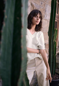 Woman in a white sweater and beige pants standing between two cacti with a stone wall background