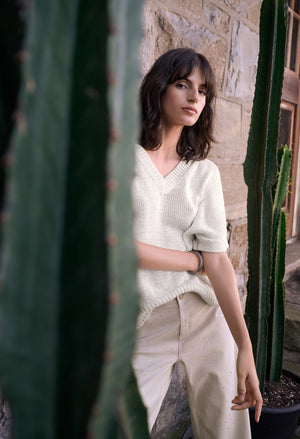 Woman in a white sweater and beige pants standing between two cacti with a stone wall background