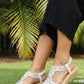White sandals with beige soles worn by a person on a stone path with greenery in the background