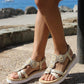 Person wearing beige sandals with decorative elements on a pebbly surface near water.