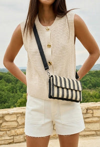 Person wearing a beige sleeveless vest and white shorts, holding a black and white striped bag with a scenic background.