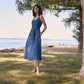 Woman in a blue dress standing in a grassy area near a body of water with trees in the background.