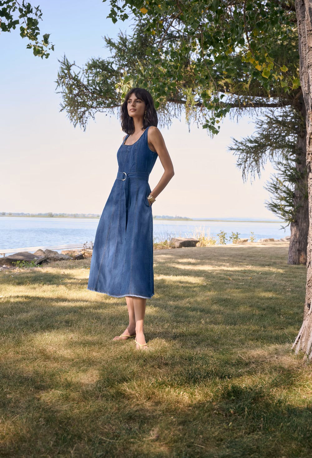 Woman in a blue dress standing in a grassy area near a body of water with trees in the background.