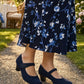 Person wearing a navy floral dress and navy shoes on a gravel path with greenery in the background