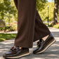 Person wearing brown dress pants and dark loafers walking on a paved path with trees and benches in the background.