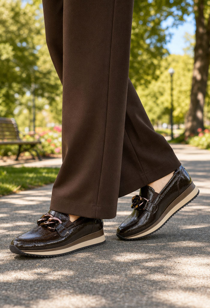 Person wearing brown dress pants and dark loafers walking on a paved path with trees and benches in the background.