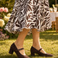 Person wearing a brown and white patterned dress with brown shoes on grass