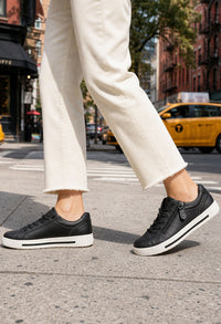Person wearing black sneakers on a city street with a yellow taxi in the background