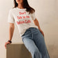 Woman wearing a white t-shirt with red text and blue jeans, leaning against a neutral background.
