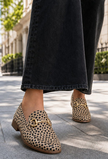 A person wearing black wide-leg jeans and leopard-print loafers with gold horsebit details, standing on a paved outdoor walkway.