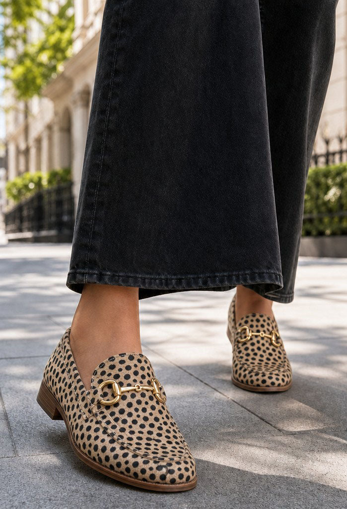A person wearing black wide-leg jeans and leopard-print loafers with gold horsebit details, standing on a paved outdoor walkway.