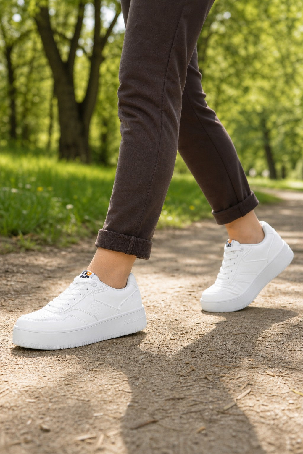 White platform sneakers worn by a person walking on a dirt path in a sunny park with green trees in the background.