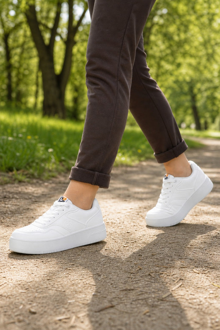 White platform sneakers worn by a person walking on a dirt path in a sunny park with green trees in the background.