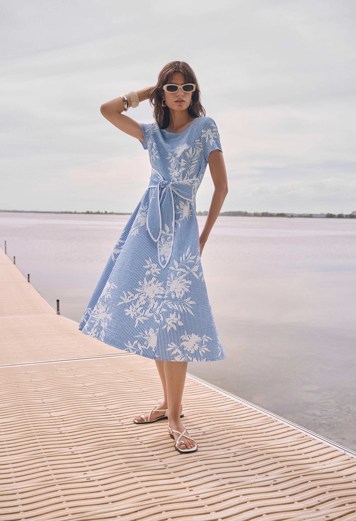 A woman wearing a light blue midi dress with a white floral pattern stands on a wooden boardwalk next to a calm body of water under an overcast sky.
