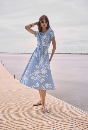 A woman wearing a light blue midi dress with a white floral pattern stands on a wooden boardwalk next to a calm body of water under an overcast sky.
