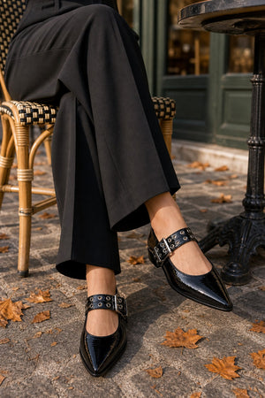 A person seated on a woven chair outdoors wearing glossy black pointed-toe Mary Jane shoes with silver buckle straps, paired with black wide-leg pants. Fallen leaves and a stone patio are visible on the ground.