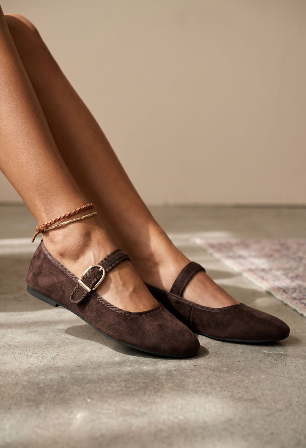 A pair of dark brown suede Mary Jane flats with a gold-tone buckle, shown on a model's feet against a neutral beige wall and concrete floor.