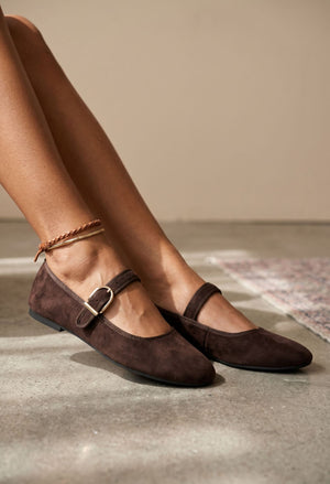 A pair of dark brown suede Mary Jane flats with a gold-tone buckle, shown on a model's feet against a neutral beige wall and concrete floor.