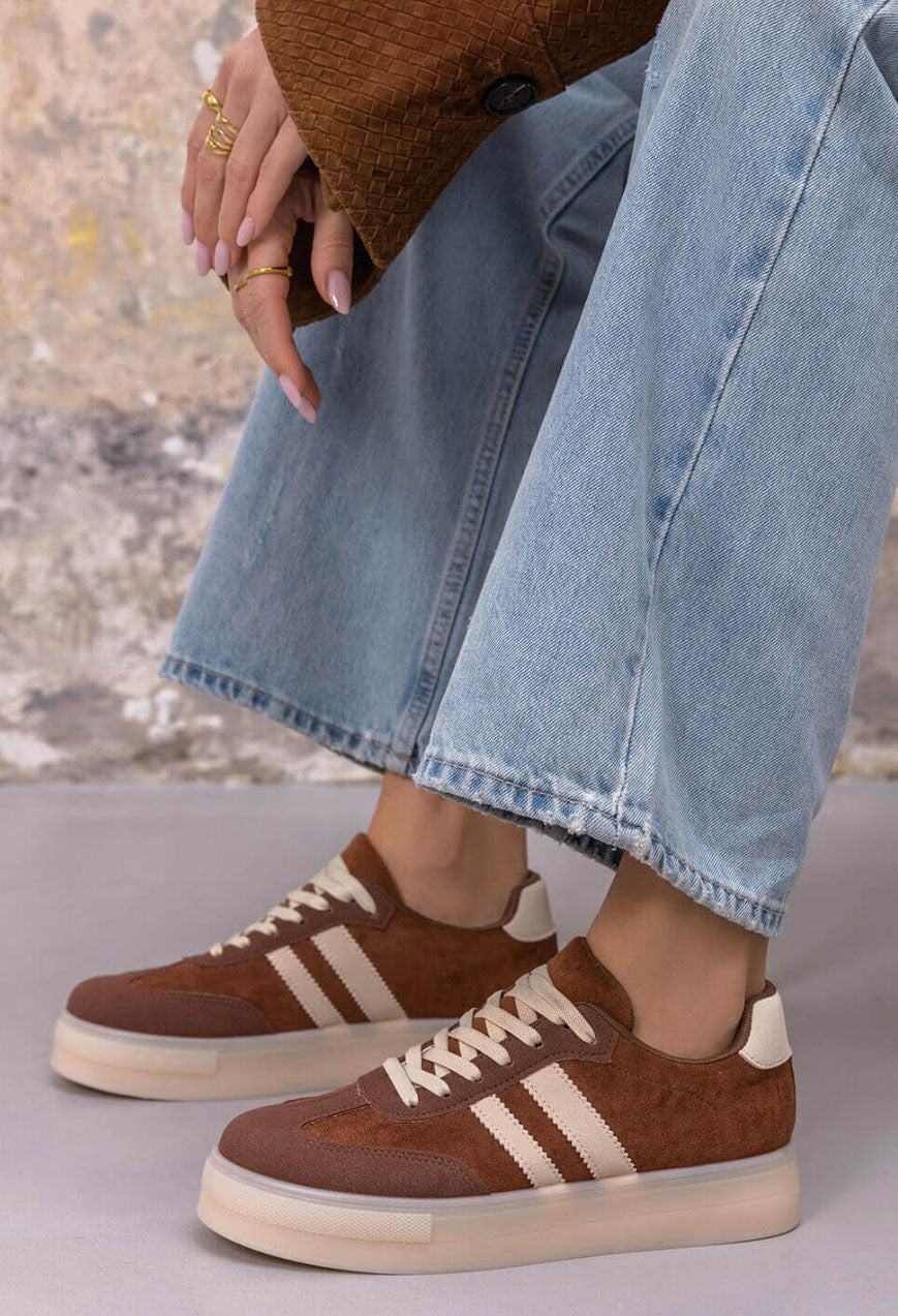A person wearing brown suede and canvas low-top sneakers with cream stripes and laces, paired with light-wash wide-leg jeans and gold rings, seated against a textured wall.