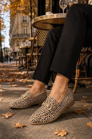 A person seated at an outdoor cafe wearing beige loafers with a black leopard print pattern and gold-tone buckle details, paired with black trousers. Fallen autumn leaves are scattered on the pavement, and a Parisian street scene is visible in the background.