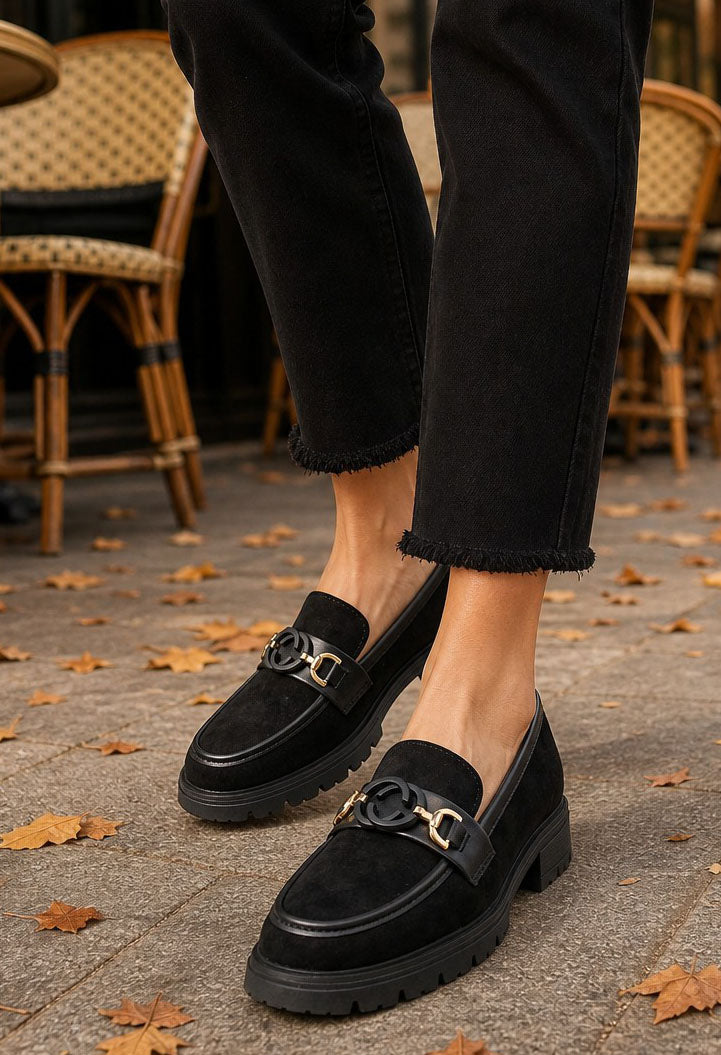 A person wearing black loafers with chunky lug soles and a decorative metal hardware strap across the vamp, paired with black cropped jeans in an outdoor cafe setting with scattered autumn leaves on the ground.