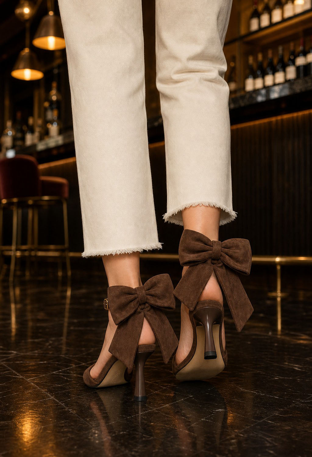 A close-up of a person's legs wearing beige cropped jeans and brown heeled sandals with large bow details, standing on a dark bar floor.