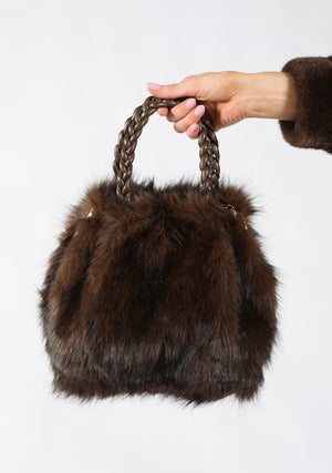 A dark brown fur handbag with a braided top handle, held by a person's hand against a plain white background.