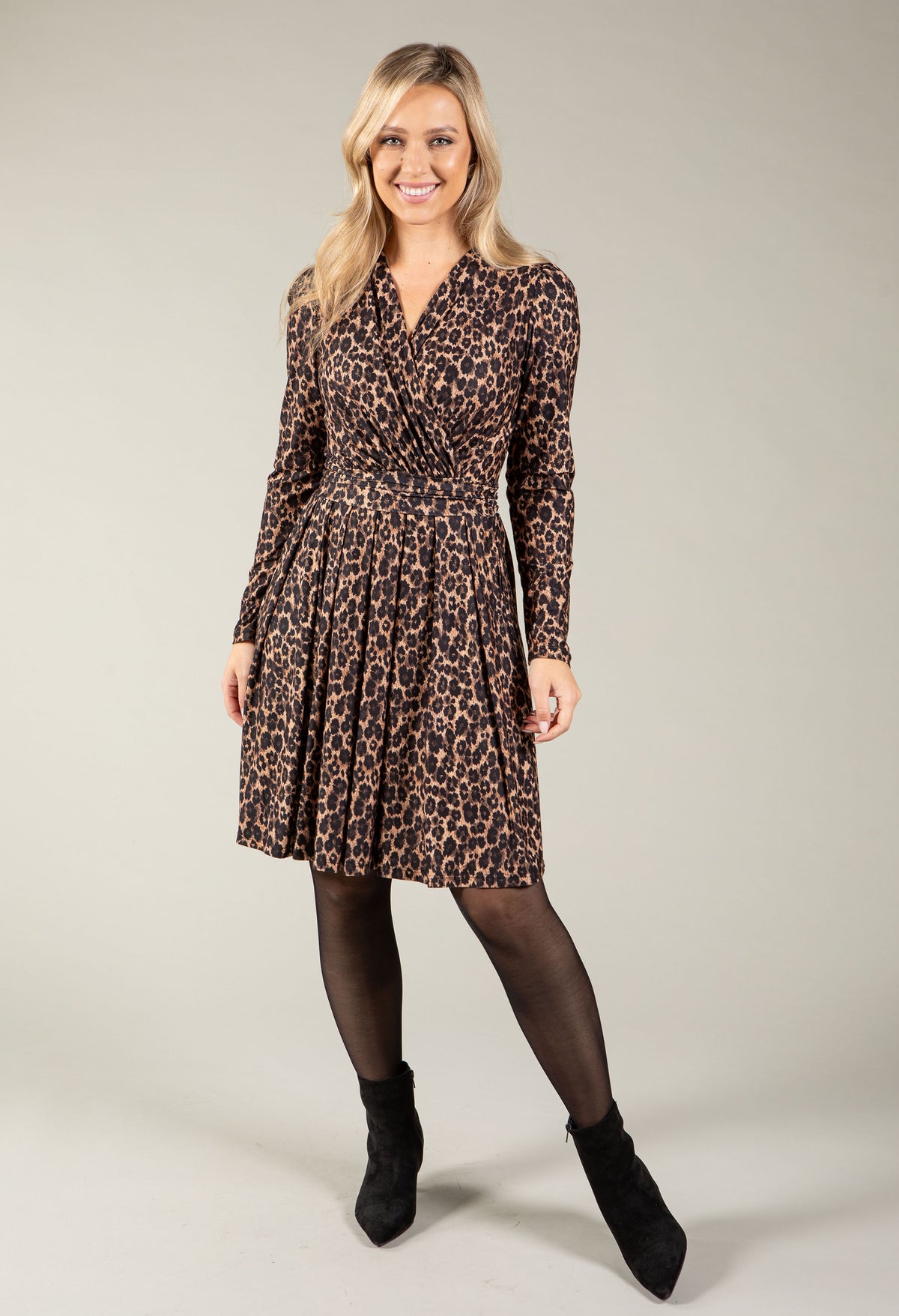 A woman modeling a long-sleeve leopard-print dress with a V-neck and flared skirt, paired with black tights and black ankle boots, against a plain light gray studio background.