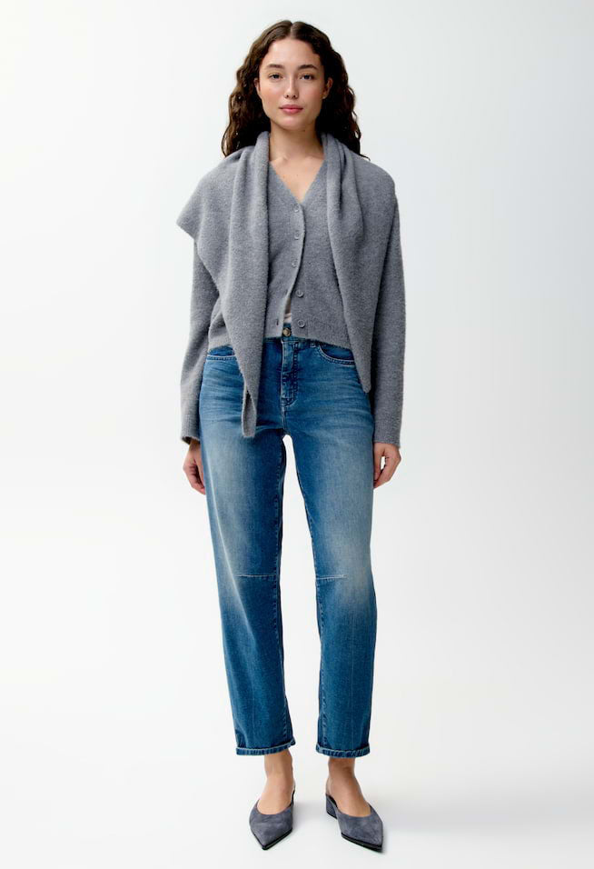 A woman modeling a gray draped cardigan over a matching gray button-front cardigan, paired with blue jeans and gray pointed flats, against a plain light gray studio background.