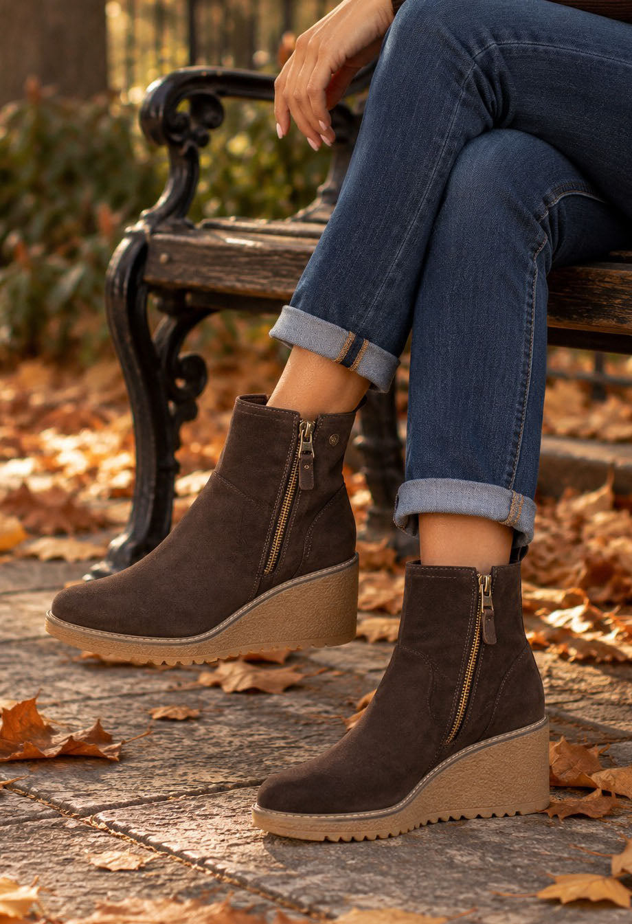 A person sitting on a park bench wearing dark brown suede wedge ankle boots with side zippers and tan soles, paired with cuffed blue jeans, with fallen autumn leaves on the ground.