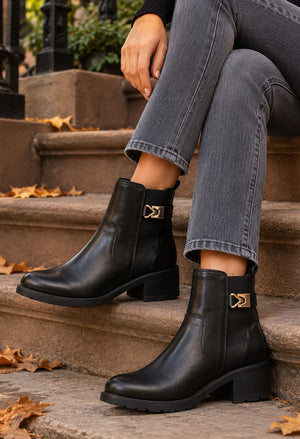 Black leather ankle boots with a gold-tone decorative buckle detail on the side, worn by a model sitting on outdoor stone steps.